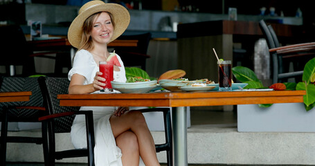 Young woman dining outdoors at cafe, wearing straw hat, enjoying refreshing smoothie. Relaxed...
