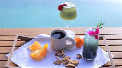 Wooden table near turquoise pool featuring refreshing breakfast spread with coffee, juice, fruits, cocktails suggesting vacation relaxation