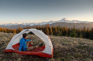 Cheerful man relaxing inside a tent, enjoying a vibrant sunset over snowy mountain peaks. Camping,...