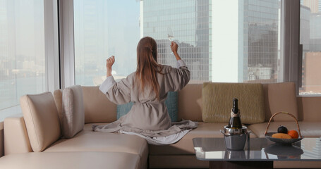 Woman in robe sitting on luxury hotel sofa with city view, enjoying champagne, relaxed, urban lifestyle, modern comfort.