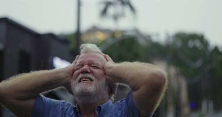 Elderly man gripping head in anguish while standing in the rain, eyes shut and expression...