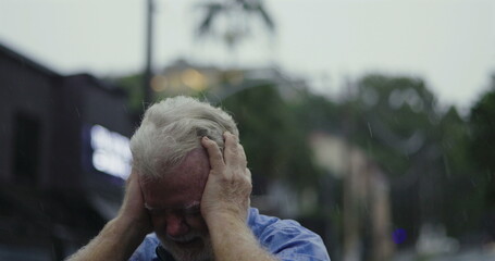 Elderly man gripping head in anguish while standing in the rain, eyes shut and expression...