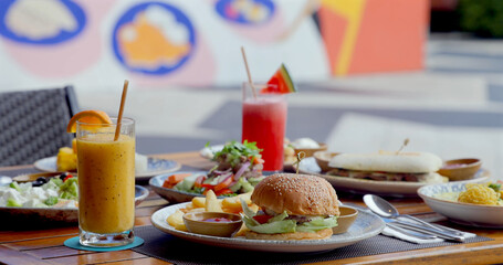 Outdoor cafe brunch includes fresh juice, burger, salad, sandwiches in vibrant urban setting. Relaxed fine dining concept with colorful background.