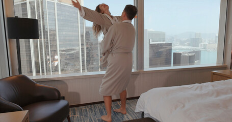 Couple in hotel room with city view, enjoying morning together in robes. Urban skyline, relaxation, romance, travel concept.