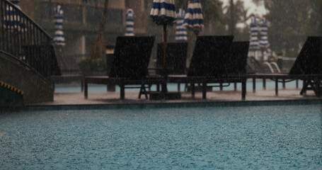 Rainy day at outdoor swimming pool with loungers and umbrellas, creating tranquil atmosphere. Captures relaxation, leisure, and nature's beauty in urban resort setting.