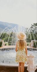 Woman in summer dress and hat standing by fountain with angel statue, mountains in background, serene outdoor scene, nature and relaxation concept.