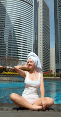 Woman in swimsuit with towel on head sitting by urban pool, skyscrapers in background, city relaxation, summer leisure, outdoor cityscape. Vertical
