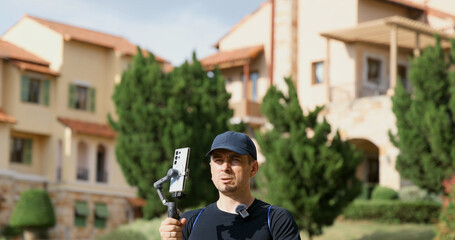 Man with gimbal camera stands in front of luxury villa in sunny estate. Filmmaking, technology, real estate concept.