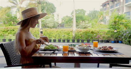 Outdoor brunch scene with woman in straw hat dining at tropical resort. Fresh salad, juice on table. Relaxing vacation, leisure.