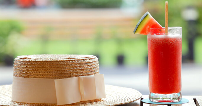 Straw hat resting on table beside refreshing watermelon smoothie outdoors in sunny park setting. Summer vibes, leisure, relaxation.