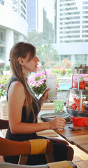 Woman enjoying refreshing beverage at outdoor restaurant in urban setting, surrounded by flowers and modern architecture, conveying relaxation, leisure, and gastronomy. Vertical