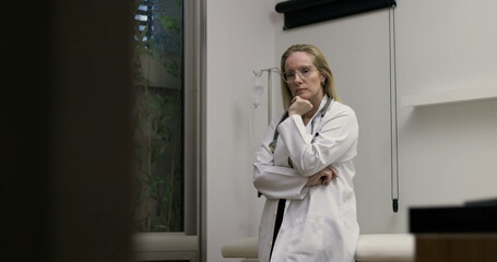 Female doctor standing in medical examination room at night, arms crossed and hand on chin in thoughtful pose, serious expression while contemplating case