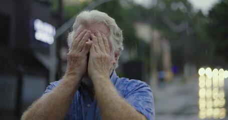 Elderly man standing still with face completely covered by hands in rain, expressing emotional...