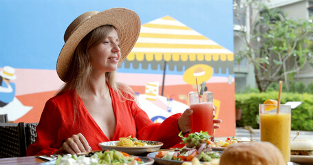 Woman wearing straw hat in outdoor cafe enjoying fresh juice and pasta. Bright mural backdrop adds vibrant summer vibe. Relaxation, dining, leisure, sunny day, outdoor eating concept.