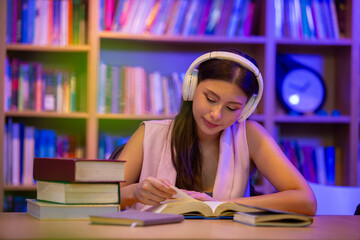 Young woman wearing headphones while reading and studying in a library at night, representing...