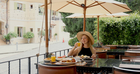 Woman enjoying breakfast outdoors at cafe, wearing straw hat, sitting under umbrella in sunny courtyard, surrounded by greenery.