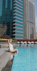 Woman relaxing by rooftop pool in urban setting, skyscraper backdrop, wearing swimsuit and hat, enjoying sunny day, cityscape relaxation. Vertical