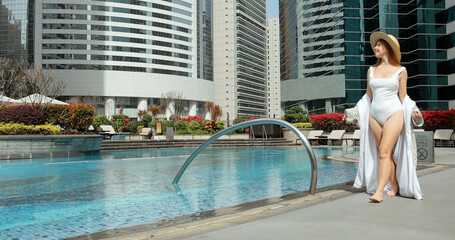 Woman in swimsuit with sunhat walks by outdoor pool at luxury urban hotel surrounded by...