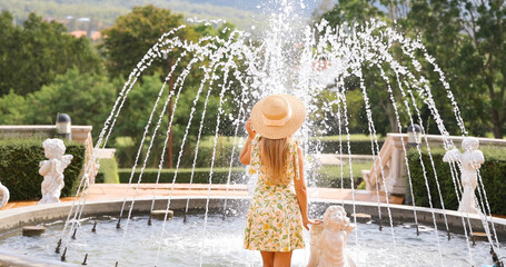 Woman in summer dress and hat enjoys serene moment in ornate garden fountain with statues, lush greenery, relaxation, elegance, and tranquility.