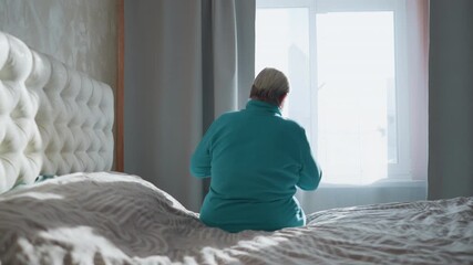 Old woman in green sitting on bed, gazing out the window. Soft morning sunlight pours in, casting a peaceful glow. She seems contemplative, enjoying the quiet solitude in her room - Powered by Adobe