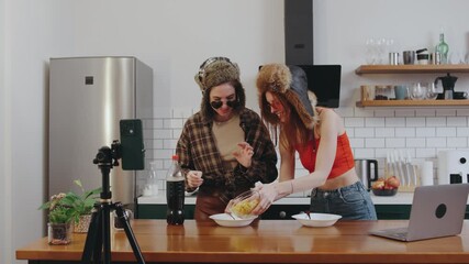 Two quirky bloggers in earflap hats and heart-shaped glasses pour soda over cereal, filming hilarious food experiments for their followers in a cozy kitchen - Powered by Adobe