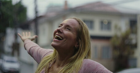 Blonde woman smiling joyfully in residential street under rain, looking up with emotional release...