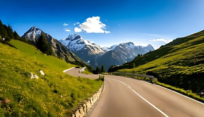 Winding mountain road curves through vibrant green hillsides, leading towards snow-capped peaks under a bright blue sky
