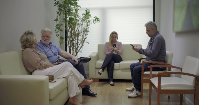 Group of patients in clinic waiting area, including elderly couple talking and others using phones, seated on couches and chairs in calm setting