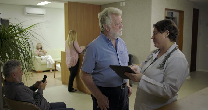 Senior man reviewing information on digital tablet with female doctor in clinic lobby, healthcare discussion in hallway with reception and patients nearby