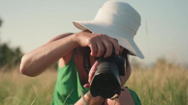 close up of woman in green dress holding camera kneeling in tall grass as she takes picture of wild plant in golden hour light with warm focus and gentle concentration on subject