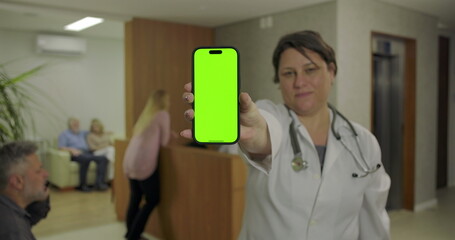 Female doctor shows phone with green screen display in hospital lobby while people wait in background at reception desk near elevator and seating area