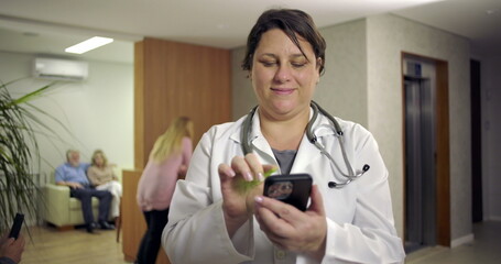 Doctor using phone with slight smile in modern hospital lobby while patients wait and nurse works...