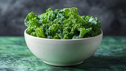 Fresh green kale leaves are arranged in a white bowl, sitting on a mottled green surface against a muted grey background, suggesting healthy eating choices.