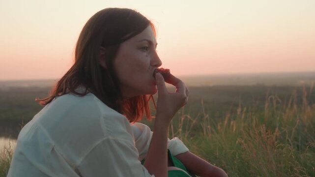 Side view of elegant lady in white shirt biting into juicy ripe plum while sunset light flares across her profile, seated in tall grassy meadow with blurred countryside