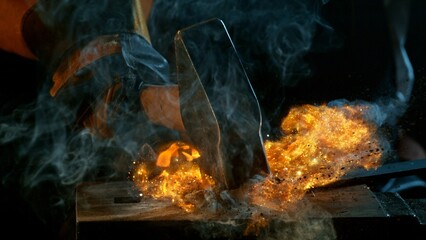 The blacksmith manually forging the molten metal on the anvil in smithy with spark fireworks