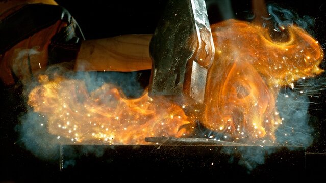 The blacksmith manually forging the molten metal on the anvil in smithy with spark fireworks