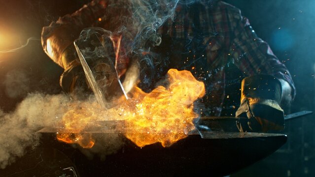 The blacksmith manually forging the molten metal on the anvil in smithy with spark fireworks