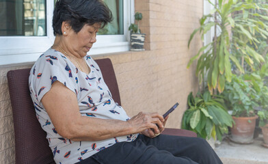 Senior woman sitting outdoors using smartphone, wearing casual clothes, surrounded by plants....