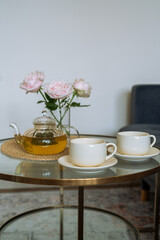 Cozy living room interior with glass teapot, two cups and pink roses in a vase on round coffee table near sofa.