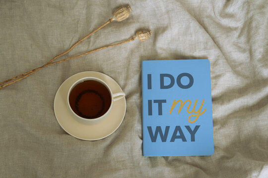 Cup of coffee or tea with blue notebook “I do it my way” on linen fabric, minimal aesthetic flat lay with warm tones.
