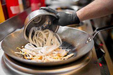 Chef pours rice noodles into wok with shrimp and bean sprouts during stir-fry preparation. Asian cuisine, kitchen action, close-up cooking scene