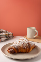 Sprinkled croissant with powdered sugar resting on a white dish on a white surface. The background is salmon pink and there are a wooden tray with a white mug and a checkered rag.