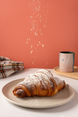 Croissant being sprinkled with powdered sugar resting on a white dish on a white surface. The background is salmon pink and there are a wooden tray with a white mug and a checkered rag.