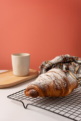 Sprinkled croissant with powdered sugar resting on a cooling rack on a white surface. The background is salmon pink and there are a wooden tray with a white mug and a checkered rag.