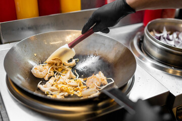 Chef stir-frying shrimp, bean sprouts, mushrooms, and egg in a wok at a professional kitchen. Close-up of cooking process with gloved hands and colorful condiments in background.