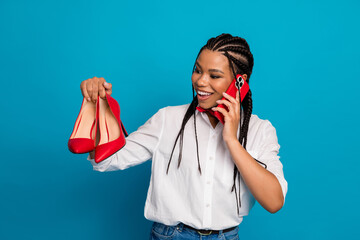 Smiling young woman holding red high heels and talking on a phone against a blue background,...