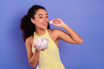 Cheerful young woman enjoying marshmallows wearing fashionable yellow outfit against a bright...