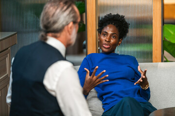 Two professionals on a sofa in a modern office, engaged in a focused conversation. One gestures while explaining, suggesting coaching, mentoring, or consulting.
