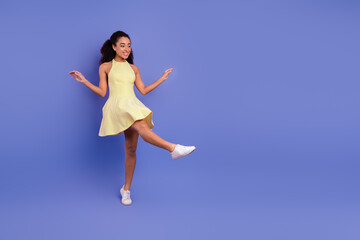 Charming young woman wearing a yellow dress smiling joyfully while dancing gracefully in front of a vibrant purple background