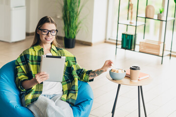 Confident woman with glasses enjoying leisure indoors while using a tablet and snacks in a modern...